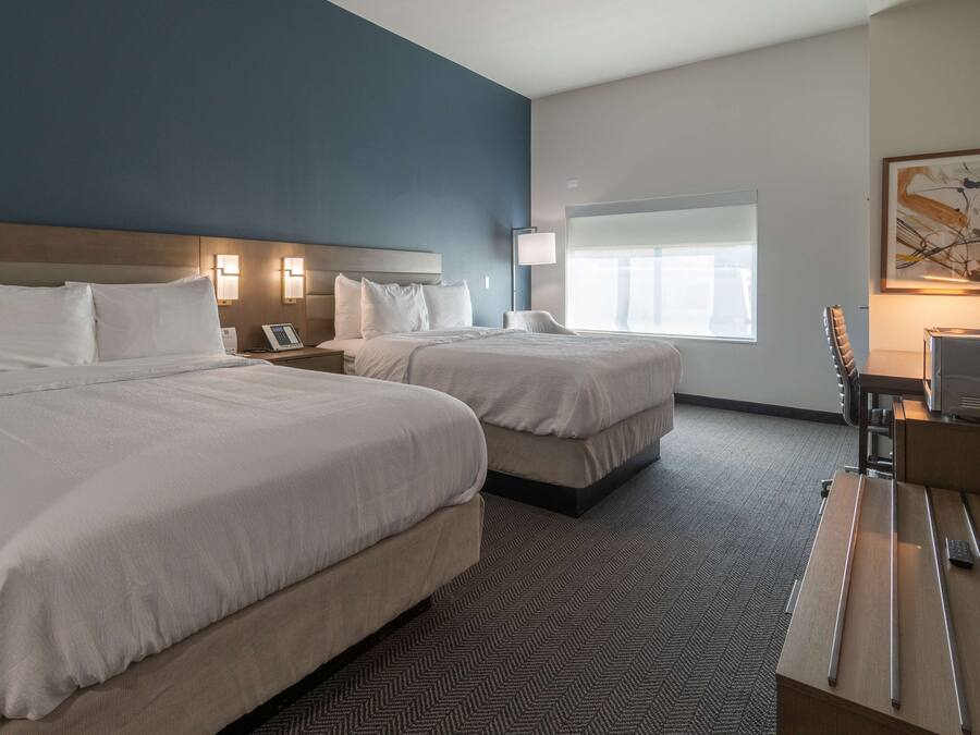 A hotel room with two beds, a desk, a lamp, a TV, and a window with blinds.