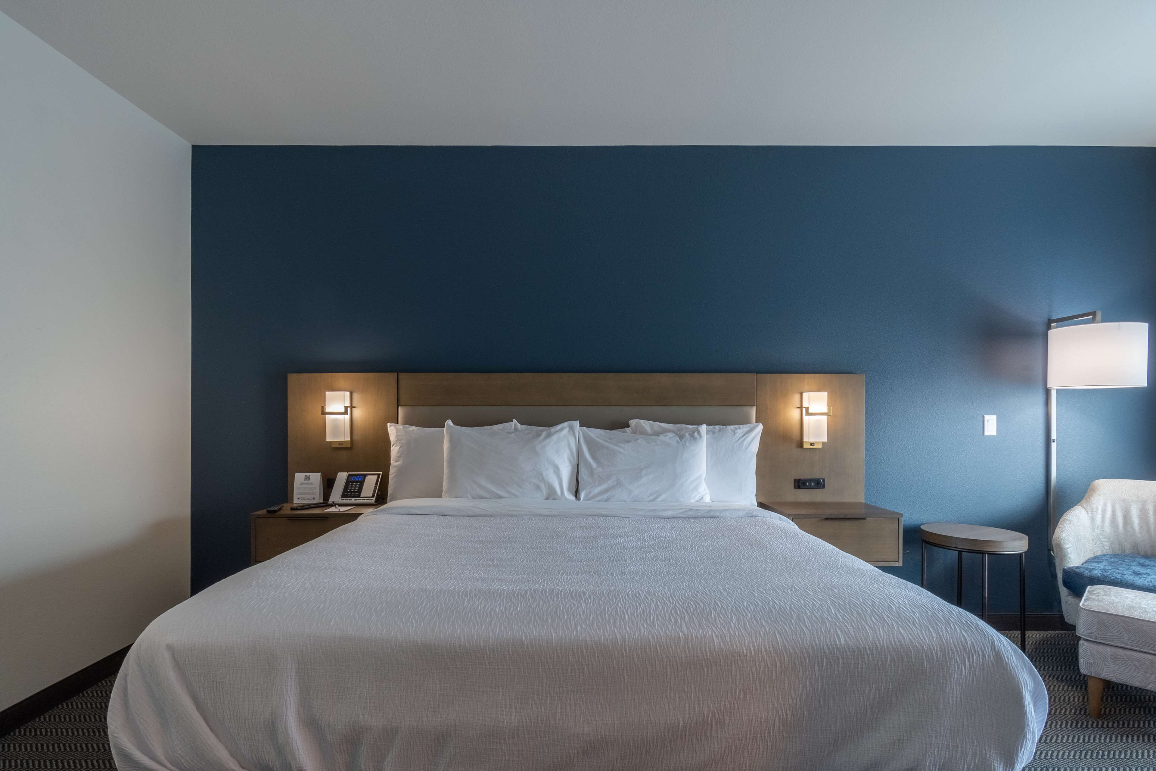 Una habitación de hotel con una cama grande, lámparas de noche, una mesita de noche, una silla y una lámpara en la pared.