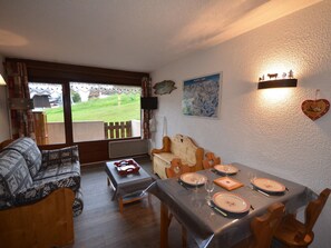 Uma sala de estar acolhedora com um sofá, um banco de madeira e uma mesa de jantar posta para quatro pessoas, com pratos e copos. Uma janela grande oferece vista para uma área gramada.