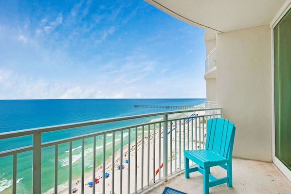 A balcony with a turquoise chair, overlooking a beach with people and umbrellas.