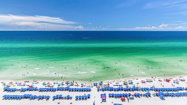 A beach with rows of blue umbrellas and sun loungers, clear turquoise water, and a clear sky.