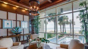 A spacious living area with a large window overlooking a pool and palm trees, featuring a wooden ceiling, comfortable seating, and framed artwork.