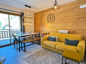 A cozy living room with a yellow sofa, a wooden wall, and a dining area with a table and chairs.