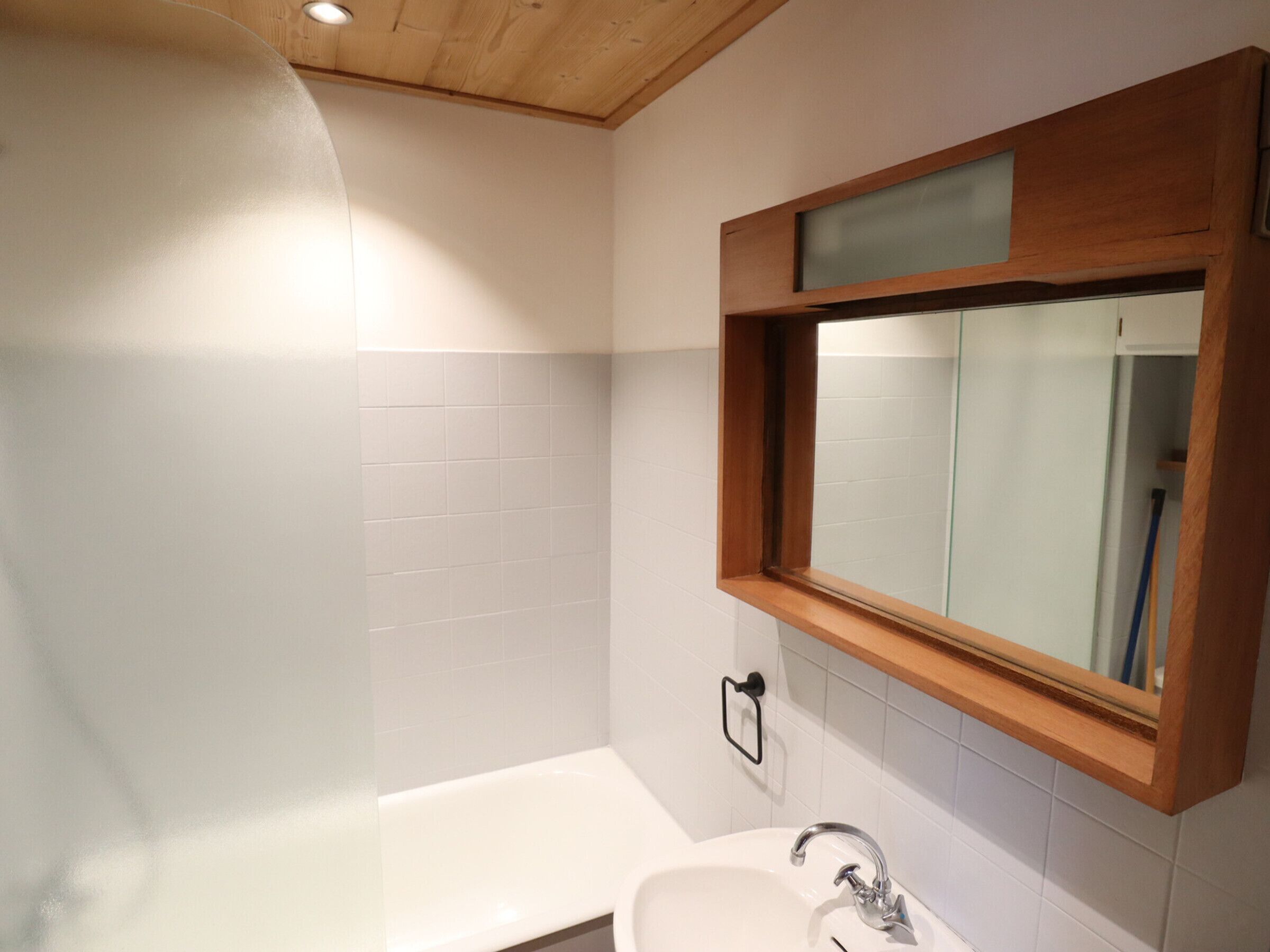 A bathroom with a white bathtub, a wooden-framed mirror, and white tiled walls.
