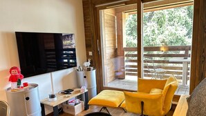 A modern living room with a TV, a small table, a yellow armchair, and a sliding glass door leading to an outdoor deck. - Modern Stylish Studio with Spa Access, Central Location, Close to Slopes (Les Gets)
