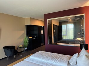 A hotel room with a large mirror, a bed with white linens, a black wardrobe, a small table with a plant, and a red accent wall.