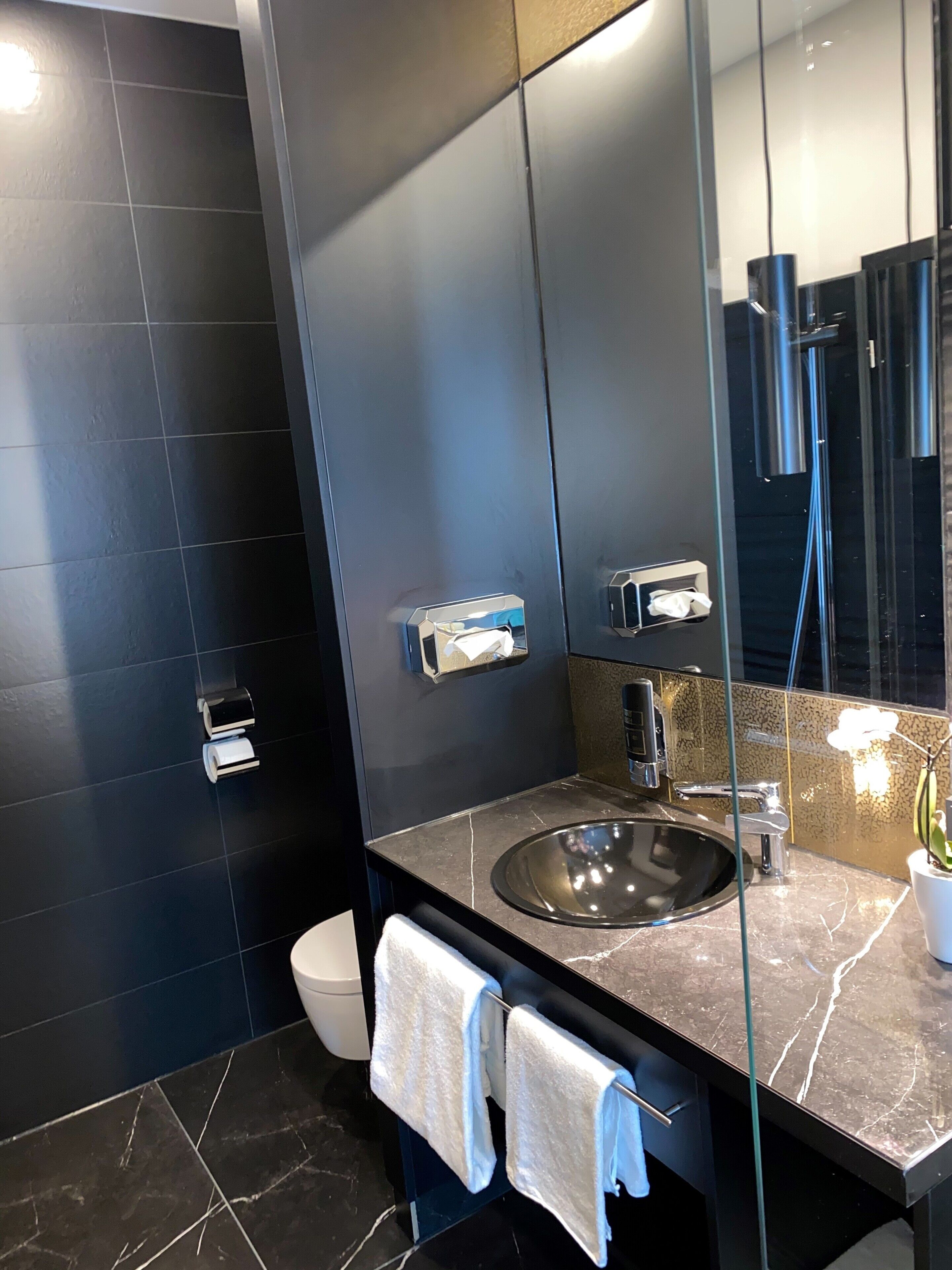 A modern bathroom with a dark tiled wall, a glass shower enclosure, a sink with a marble countertop, and two towels hanging on a rack.