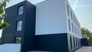 Un edificio moderno con fachada blanco y negro, numerosas ventanas y un área de acceso pavimentada.