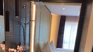 Un baño moderno con ducha de vidrio, un lavamanos con un florero y una cama visible a través del vidrio.
