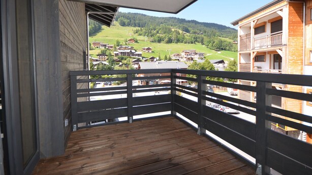 Un balcón con piso de madera y baranda de metal, con vista a un pueblo con edificios y colinas verdes.