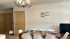 Un comedor con una mesa de madera dispuesta para cuatro, sillas blancas, una araña y un cuadro en la pared.