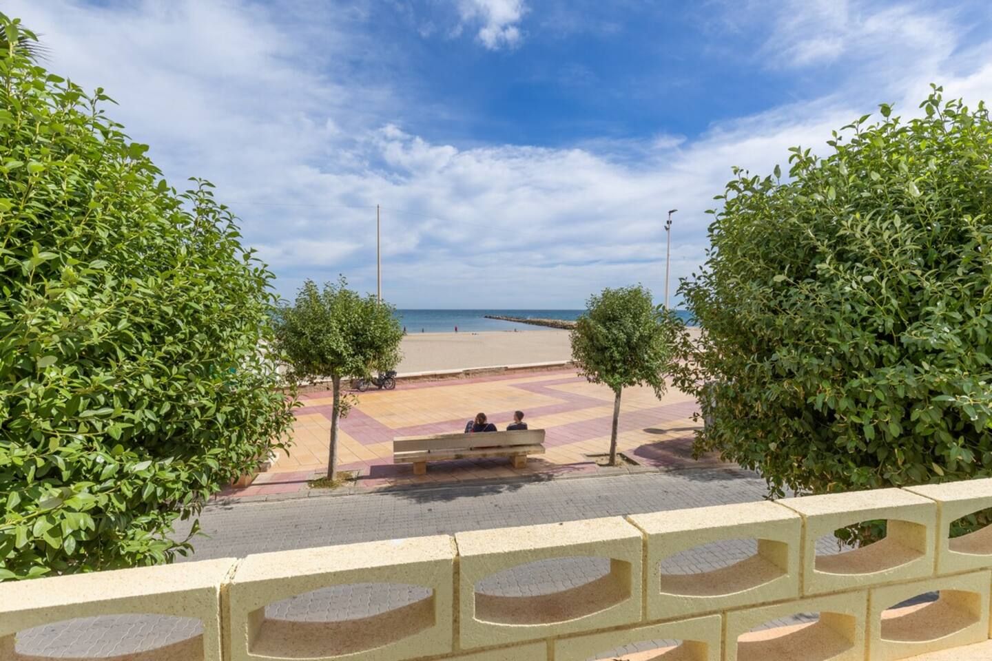 Ein gepflasterter Weg führt zu einem Strand mit einer Bank und Bäumen.