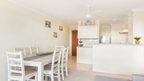 Uma sala de jantar com mesa de madeira e cadeiras brancas, uma cozinha com armários brancos e uma geladeira, e um ventilador de teto.