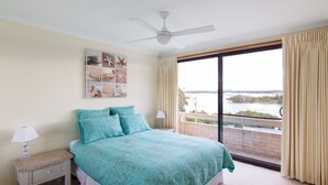 Quarto com cama, ventoinha no teto, mesa de cabeceira, abajur, janela com cortinas e vista para a água.