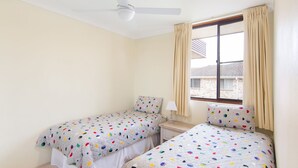 Quarto com duas camas de solteiro, ventilador de teto, janela com cortinas e vista para o exterior.