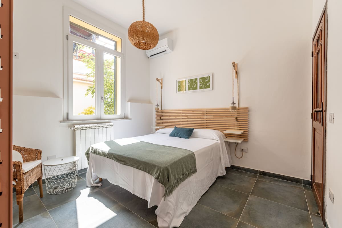 A bedroom with a bed, a window, a radiator, a wicker chair, a bedside table, and a wall-mounted lamp.