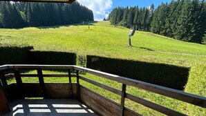 A wooden deck with a metal railing overlooking a green field with a signpost and trees in the background. - Ski-In/Ski-Out Apartment in Les Gets - 3 Stars, Balcony, Mountain Views, Free Shuttle (Les Gets)