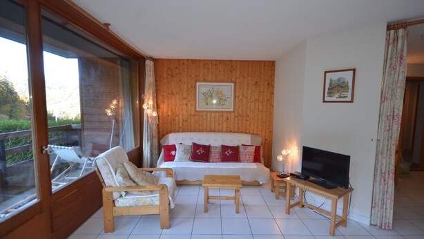 A bedroom with a bed, a TV, a wooden chair, a wooden table, and a balcony with a chair. - Ski-In/Ski-Out Apartment in Les Gets - 3 Stars, Balcony, Mountain Views, Free Shuttle (Les Gets)