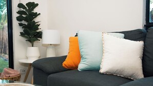 Un sofá gris oscuro con tres almohadas anaranjadas, azul claro y blancas, una lámpara blanca y una planta verde grande sobre una mesita de noche.