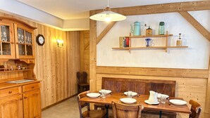 A wooden dining area with a table set for four, surrounded by chairs. There is a wooden cabinet with glass doors to the left, and a shelf with decorative items above the table.
