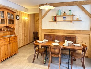 Uma sala de jantar de madeira com uma mesa posta para quatro pessoas, rodeada por cadeiras. À esquerda, um armário de madeira com portas de vidro, e acima da mesa, uma prateleira com objetos decorativos.
