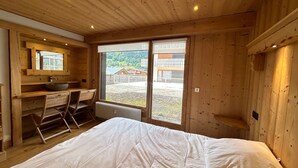 A wooden cabin room with a bed, a desk with a sink, and a large window offering a view of a building and landscape.
