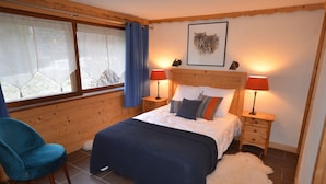 Un dormitorio con cabecera de madera, cama con ropa de cama blanca y a rayas, dos lámparas de noche, una silla, una alfombra de piel y una ventana con cortinas.