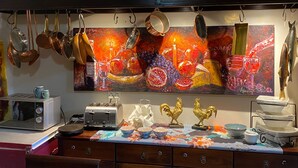 Una cocina con utensilios de cobre, un cuadro de frutas y copas de vino, un tostador y dos gallos dorados sobre la encimera.