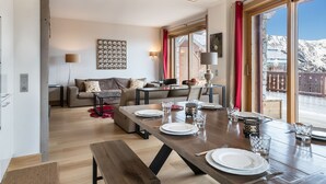 A modern living room with a dining area, wooden flooring, a sofa, a table set for four, and large windows offering a view of a snowy landscape. - Luxury Apartment, 3 Bedrooms, Close to Slopes, Sauna (Méribel)
