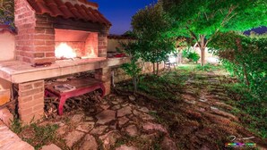 An outdoor brick oven with a fire burning inside, surrounded by a garden with trees and a stone pathway. - 4 Bed Villa Sleeps 9 - Pool, Pets, Garden, Parking (Supetar)