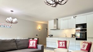 Un salon moderne avec un canapé gris, une table basse en bois et deux fauteuils dotés de coussins rouges. Un lustre est suspendu au plafond, et on aperçoit au fond une cuisine avec des meubles blancs et un four encastré.