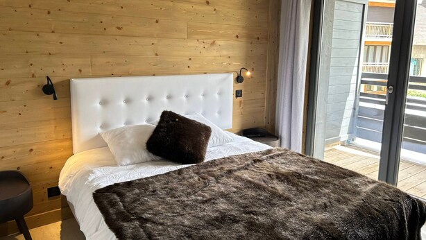 Une chambre avec un mur en bois, une tête de lit blanche capitonnée, un grand lit avec une literie blanche et marron foncé, une petite table de chevet et une commode.