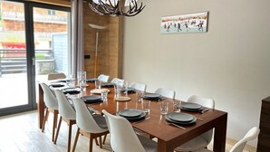 Un coin repas avec une table en bois dressée pour 10 personnes, des chaises blanches et un lustre.