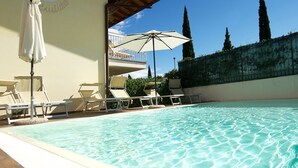 Piscina con sillones de descanso y sombrillas, un área cubierta y vistas al cielo y árboles.