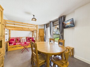 A cozy room with a wooden bunk bed, a dining table with chairs, a TV mounted on the wall, and a window with curtains. - Central Les Gets Apartment with Terrace, Garage and WIFI (Les Gets)