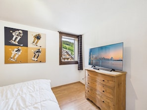 A bedroom with a bed, a wooden dresser, a TV on a wooden stand, and a cityscape painting on the wall.