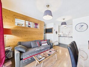 Ein gemütliches Wohnzimmer mit einer Holzwand, einem grauen Sofa, einem kleinen Couchtisch und einer Küche im Hintergrund.