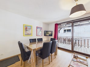 A dining area with a wooden table and chairs, a TV on the wall, and a balcony with a view.