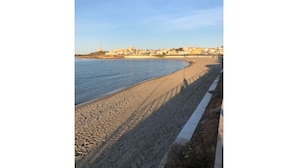 Ein Strand mit blauem Himmel, sandigem Ufer und Gebäuden im Hintergrund.