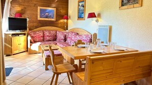 A cozy room with a wooden sofa, a dining table set for two, a television, and a red lamp. - Ski-in/Ski-out Apartment with Terrace & Summer BBQ, Near Village Center (Les Gets)