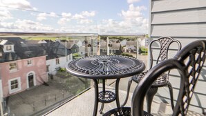 Un balcon avec une table ronde en métal et deux chaises, donnant sur un quartier résidentiel avec des maisons et un ciel dégagé.