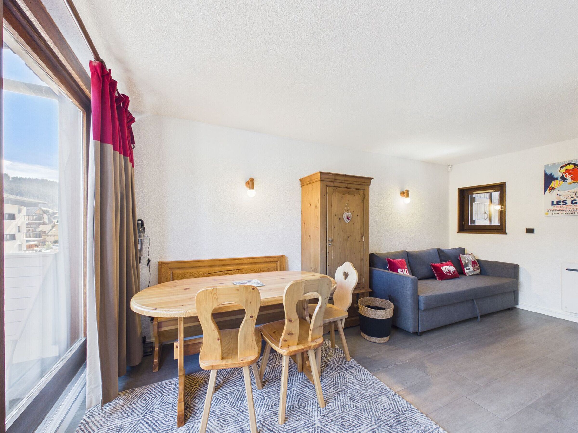 A cozy living room with a wooden dining table, two chairs, a sofa with cushions, a cabinet, and a window with curtains.