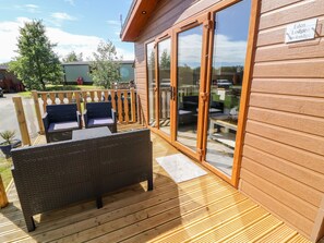 A wooden deck with wicker furniture, a glass door entrance, and a sign indicating the property name. - Eden Lodge - Sherwood 21 (Carnforth)