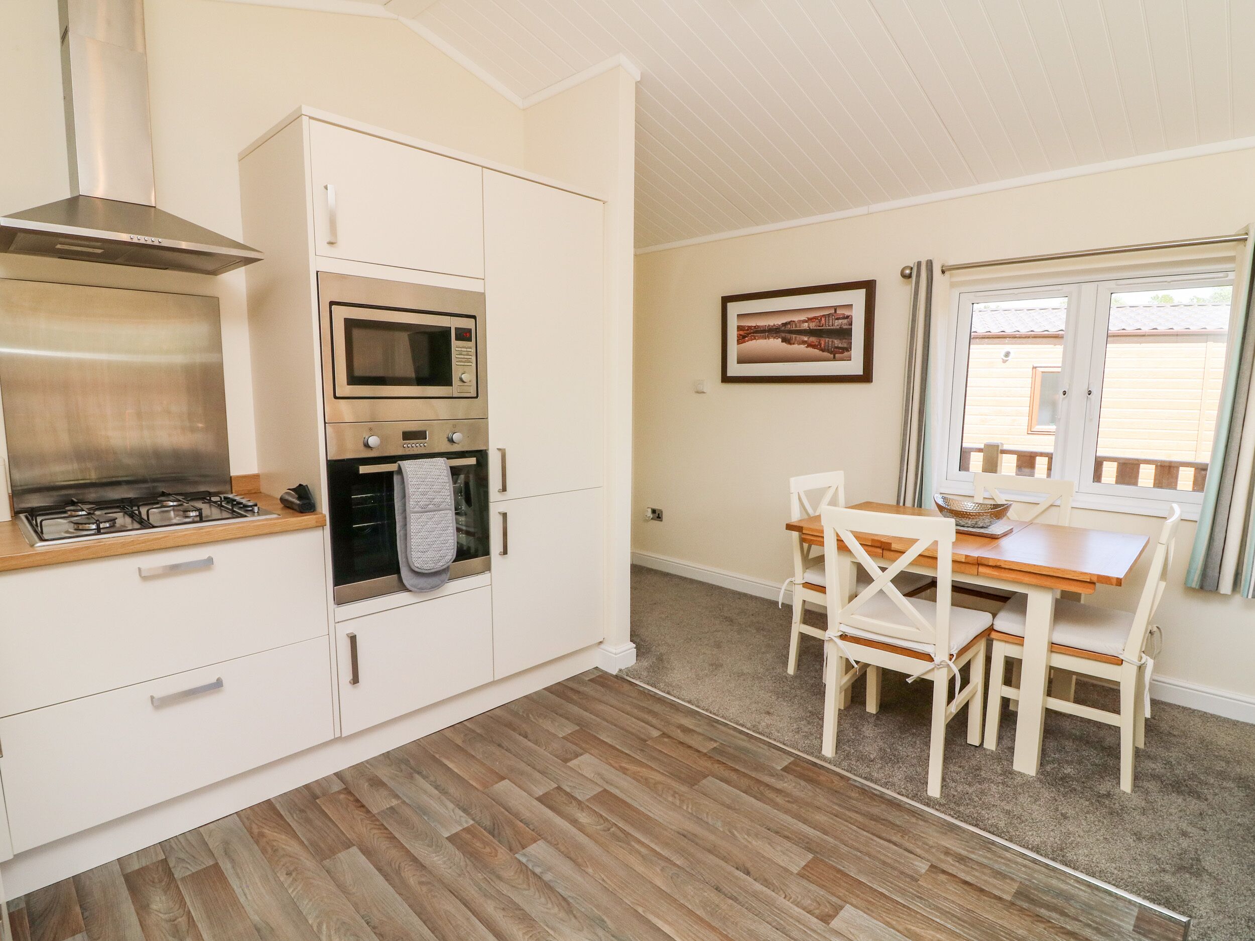 A compact kitchen with white cabinetry, a built-in oven, microwave, and gas stove. There is a dining table with white chairs and a window with curtains.