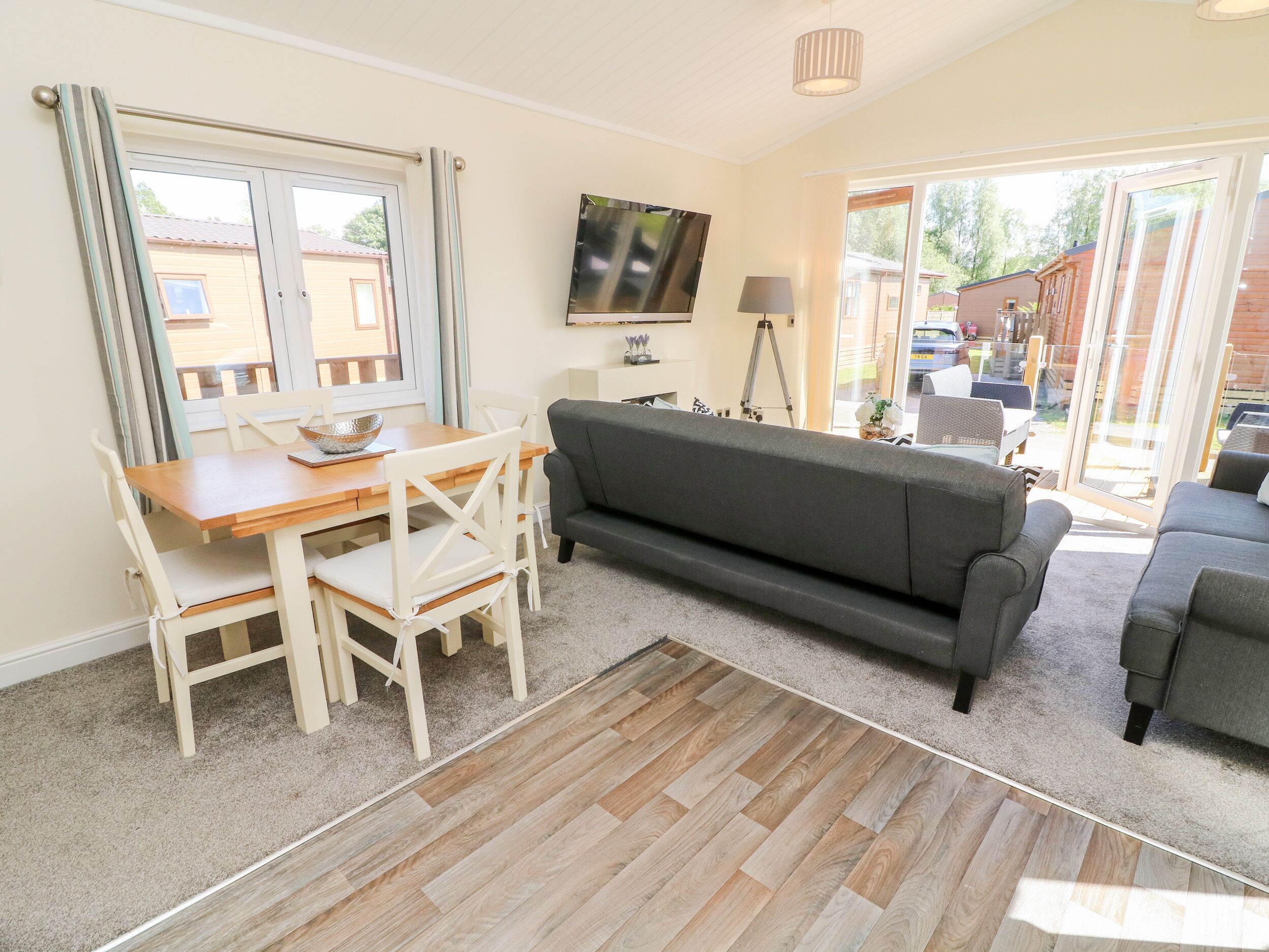 A living room with a wooden dining table, white chairs, a grey sofa, and a TV mounted on the wall.