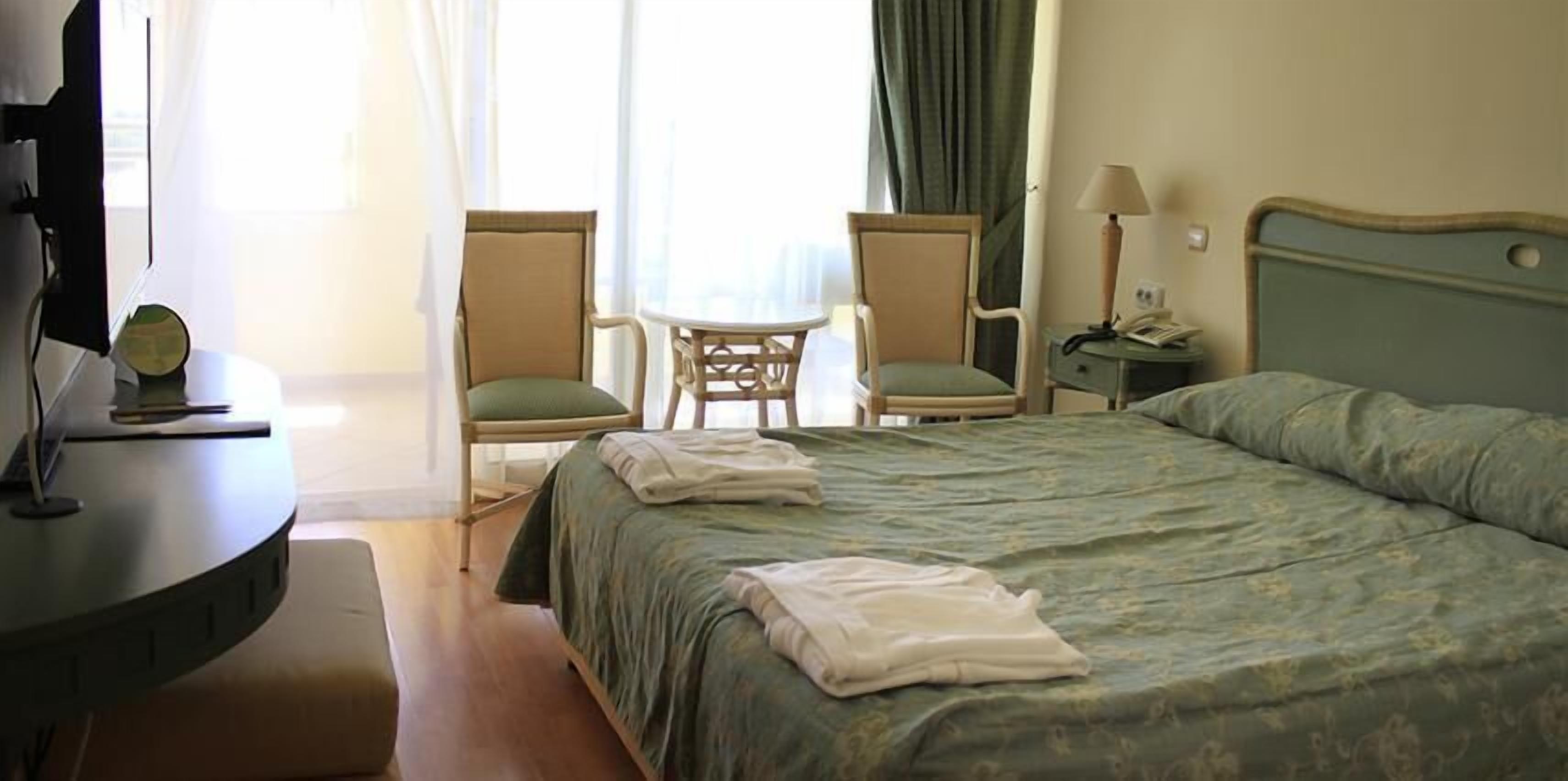 Ein Hotelzimmer mit einem Bett, zwei Stühlen, einem kleinen Tisch, einer Lampe und einem Schreibtisch mit einem Computer.
