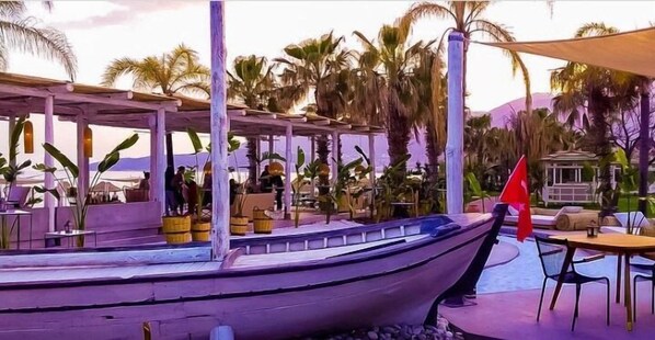 A boat docked at a waterfront restaurant with outdoor seating and palm trees in the background.