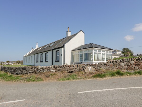 A two-story house with a stone wall in front, a clear sky, and a road in the foreground. - Ailsa Shores (Girvan)