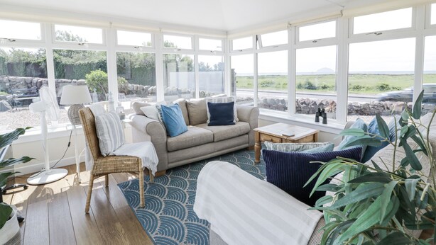 A sunlit room with a sofa, a chair, and a table. Large windows offer a view of a stone wall and greenery outside. - Ailsa Shores (Girvan)