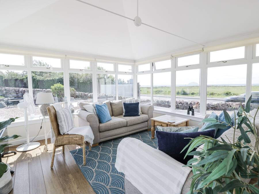 A sunlit room with a sofa, a chair, and a table. Large windows offer a view of a stone wall and greenery outside.